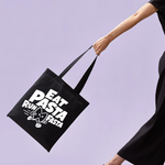 Load image into Gallery viewer, A black cotto tote bag with bold white text reading 'EAT PASTA RUN PASTA' and a small cartoon pasta illustration, carried by a person whose arm and black dress are visible against a solid light lavender background.