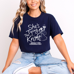Load image into Gallery viewer, A woman wearing a navy blue graphic t-shirt with white text that reads 'She's Tying The Knight' and 'ABIGAIL'S HEY FUENGIOLA 2027', paired with light-wash distressed jeans.