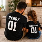 Load image into Gallery viewer, Man and child sitting on a carpeted floor wearing matching black t-shirts with 'DADDY 01' and 'DADDY'S GIRL 01' text.