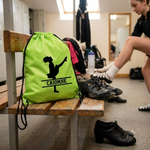 Load image into Gallery viewer, A bright lime green drawstring bag with a black silhouette of a dancer and the name 'CAIDMHE' printed on the front, sitting on a wooden bench in a locker room. A person is visible in the background adjusting their shoe.