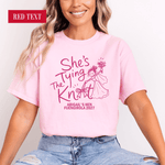 Load image into Gallery viewer, A woman wearing a light pink graphic T-shirt with dark pink text reading 'She's Tying The Knot' and a line-art illustration of a woman in a dress, paired with light blue jeans. A red label in the top-left corner says 'RED TEXT'.