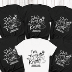 Load image into Gallery viewer, Six black and one white short-sleeve T-shirt laid flat on a whitewashed wood background, each printed with a black graphic and text. The shirts feature a bride and groom illustration and phrases including 'She's The Tying The Knot' and 'I'm Tying The Knot,' with 'ABIGAIL'S KEN FUENGIROLA 2027' printed below.
