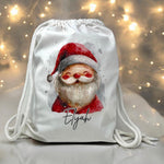 Load image into Gallery viewer, Personalised white drawstring bag with a watercolour Santa design and example name Eliyah, in front of fairy lights