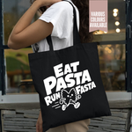 Load image into Gallery viewer, A person wearing a white t-shirt carries a black canvas tote bag with a bold white graphic that reads 'EAT PASTA RUN FASTA' and a cartoon pasta character. The photo is taken in front of a storefront window, with a pink overlay in the top right corner reading 'VARIOUS COLOURS AVAILABLE'.
