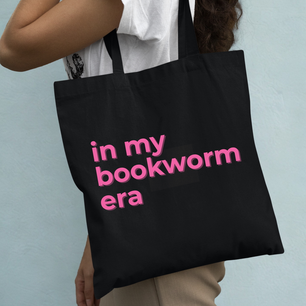 Black tote bag with pink text 'in my bookworm era' held by a person against a light blue background