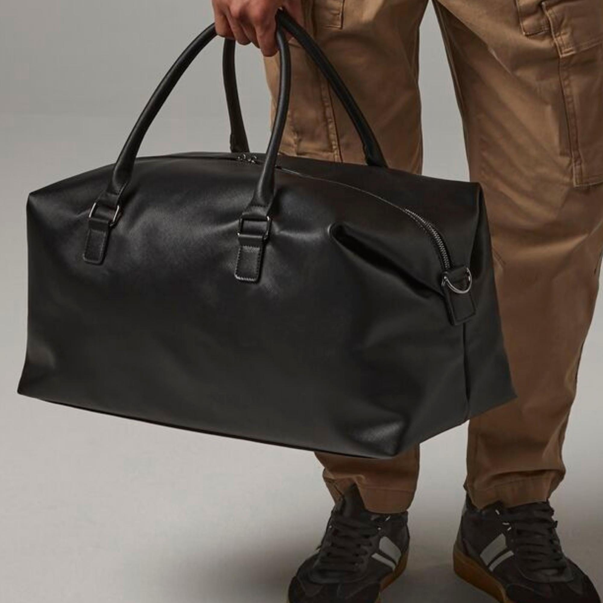 A person is holding a black leather duffel bag with a zipper closure.