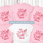 Load image into Gallery viewer, Six short-sleeve crew-neck T-shirts arranged in a collage on a whitewashed wood background, showing two color options: light pink and white. Both shirts feature a red graphic of a woman tying a knot with floral accents, with text reading 'She's Tying The Knot' and 'ABIGAIL'S HEN FUENGIROLA 2027'.

