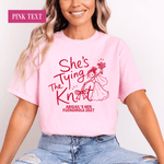 Load image into Gallery viewer, A woman wearing a light pink graphic T-shirt with red text reading 'She's Tying The Knot' and a red illustration of a woman knitting, paired with light blue jeans. A pink label in the top left says 'PINK TEXT'.
