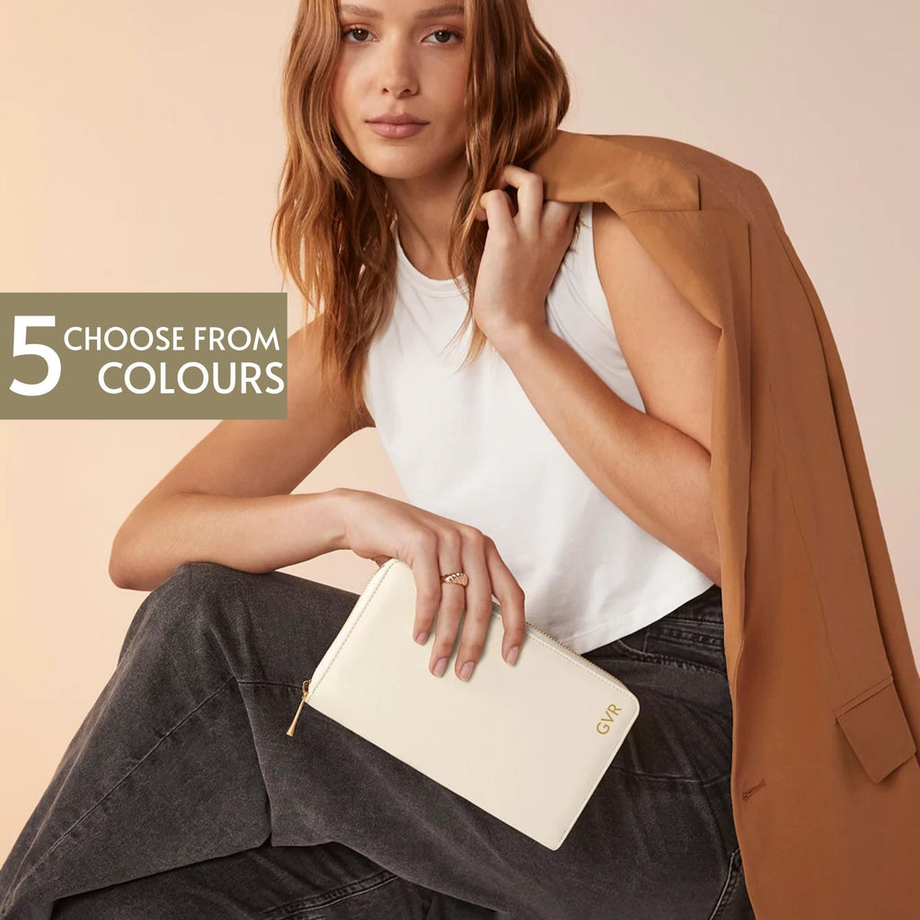 A woman with long brown hair is sitting and holding a white wallet with a gold zipper. She is wearing a white top and a brown jacket.